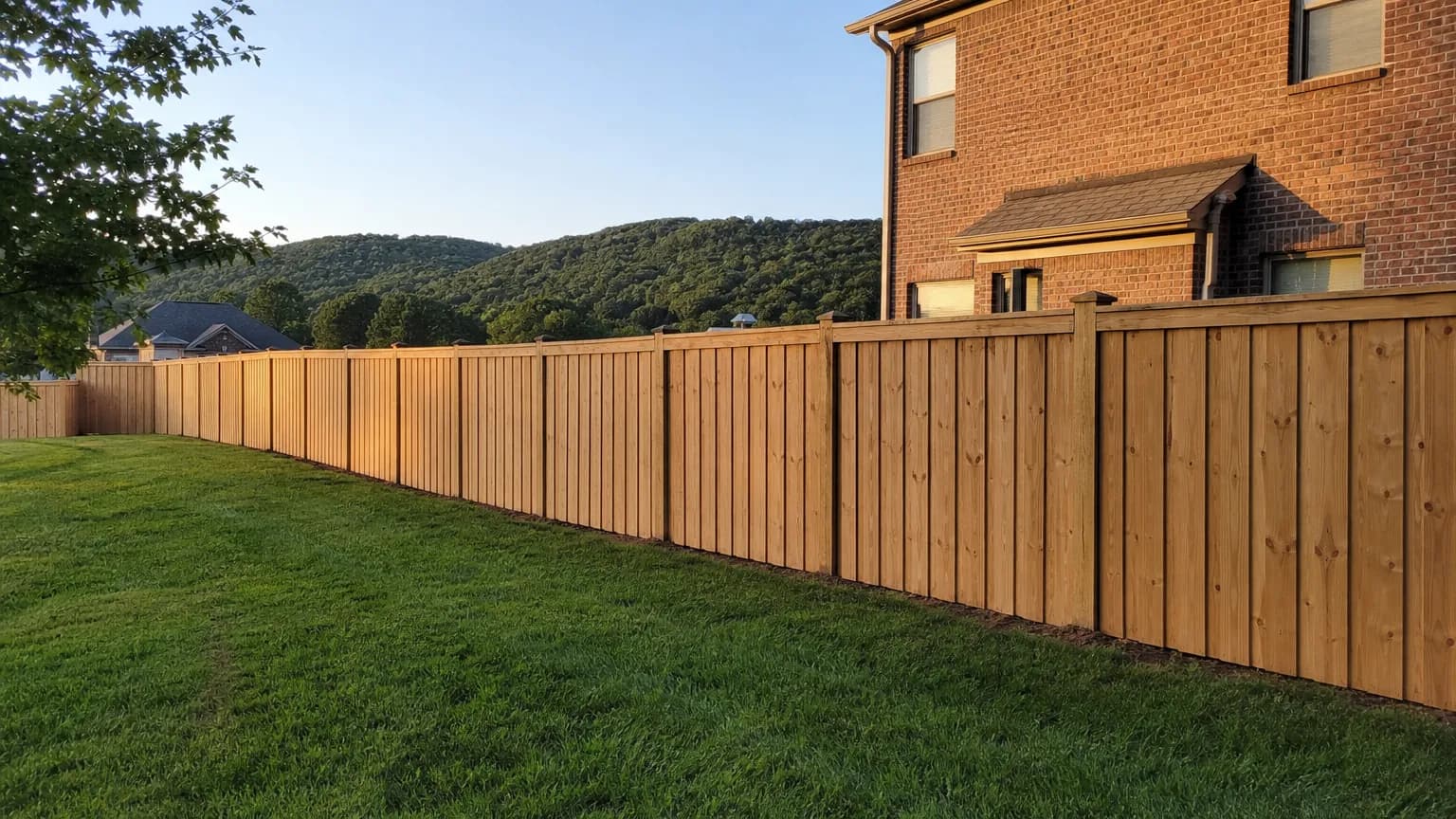 New cedar board-on-board privacy fence with a cap rail along the back yard of a brick two-story home, with a wooded ridge behind